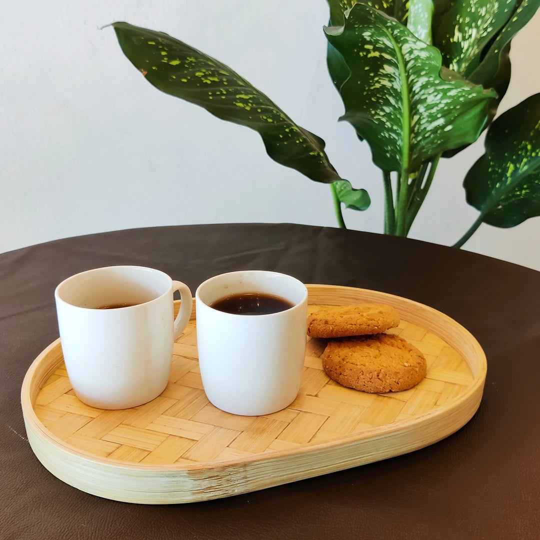 A bamboo oval serving tray with coffee and cookies, placed on a dark brown table with green plant leaves in the background.