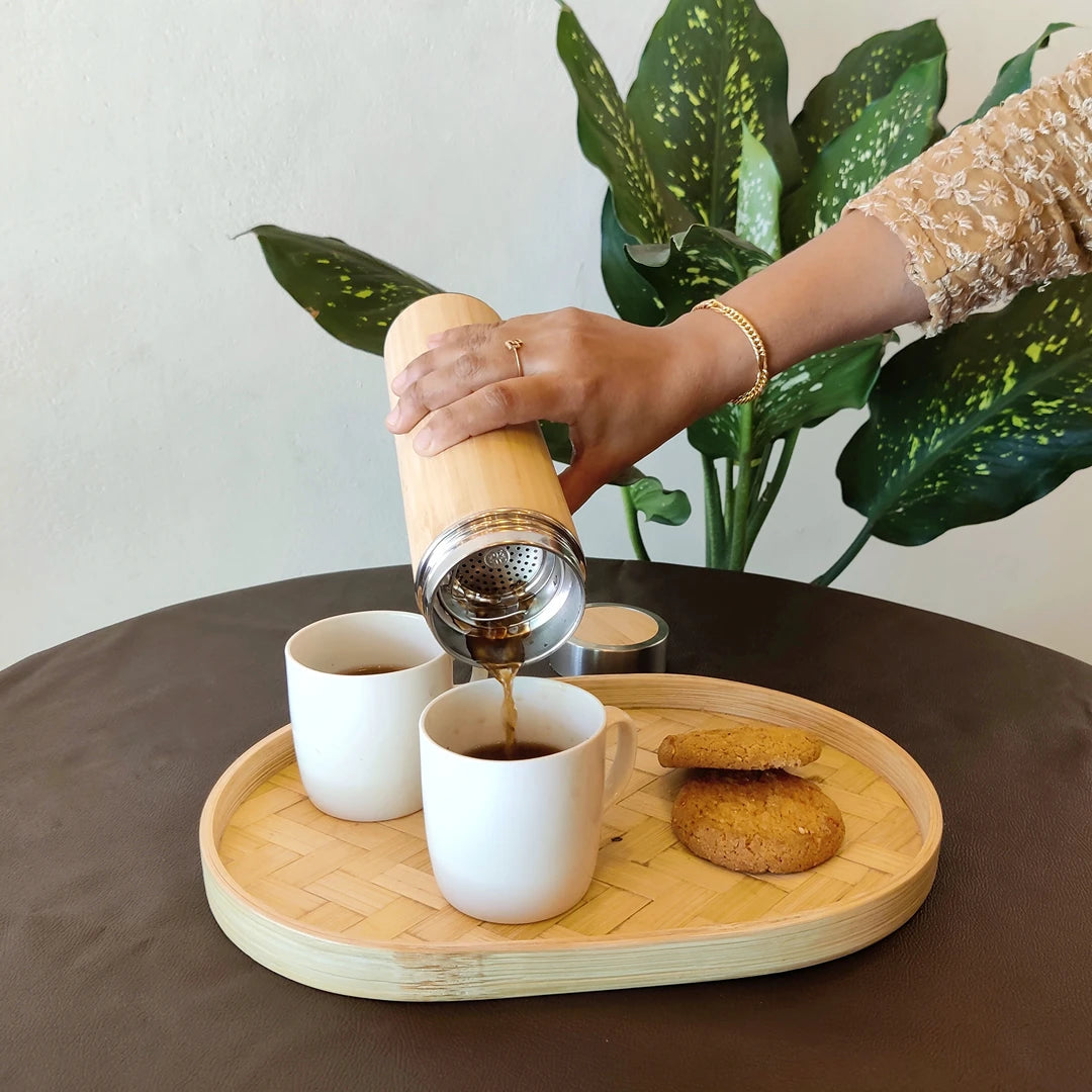 A hand pouring coffee into white mugs placed on a bamboo oval serving tray, with cookies on the side.