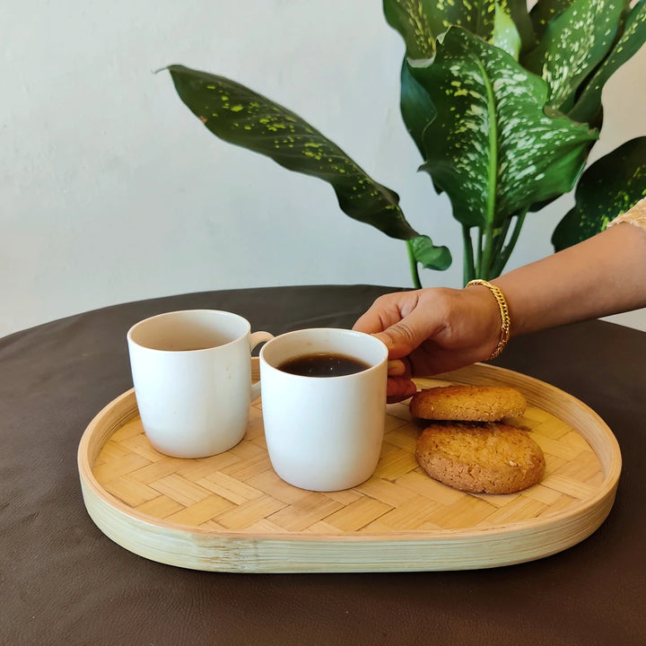 A bamboo oval serving tray with two white mugs of coffee and cookies, placed on a dark table with a plant in the background.